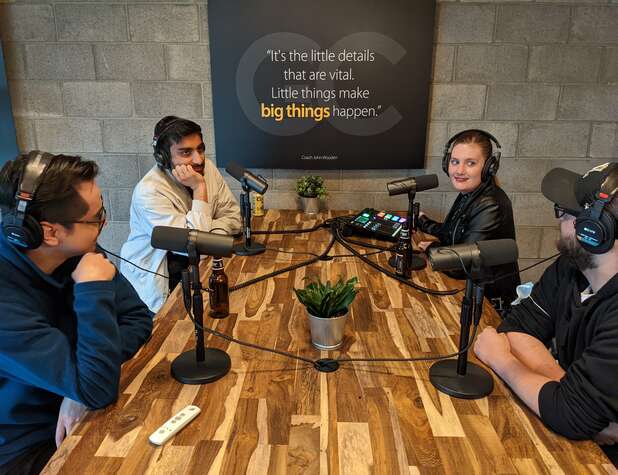 Group of 4 people around a table recording a podcast. They have headphones and are speaking into a microphone.